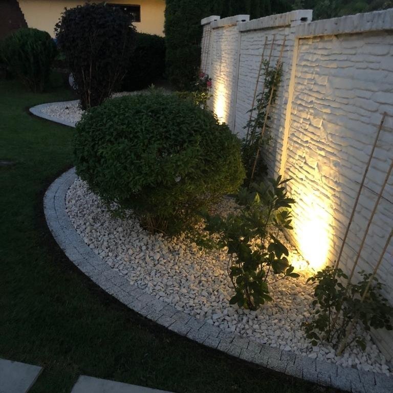 Abendlich beleuchteter Garten mit gebogenem Kiesstreifen. Darin steht ein kugelrunder Busch vor einer weißen Steinmauer, die von warmem Licht angestrahlt wird.